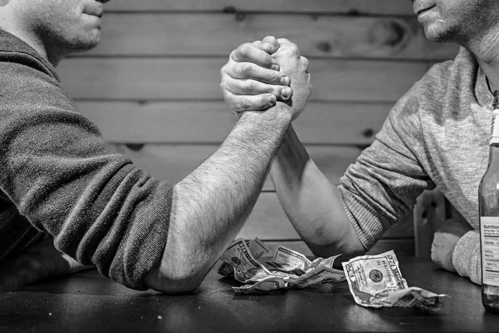 Photographie en noir et blanc de deux hommes se livrant à un bras de fer au-dessus de billets de banque. Cette photo illustre symboliquement la tension concernant les impayés rencontrés par les travailleurs indépendants.